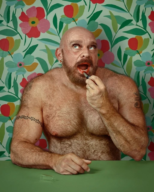 A shirtless, bald adult male with a beard applies red lipstick in front of a colorful floral wallpaper and green table, looking up.